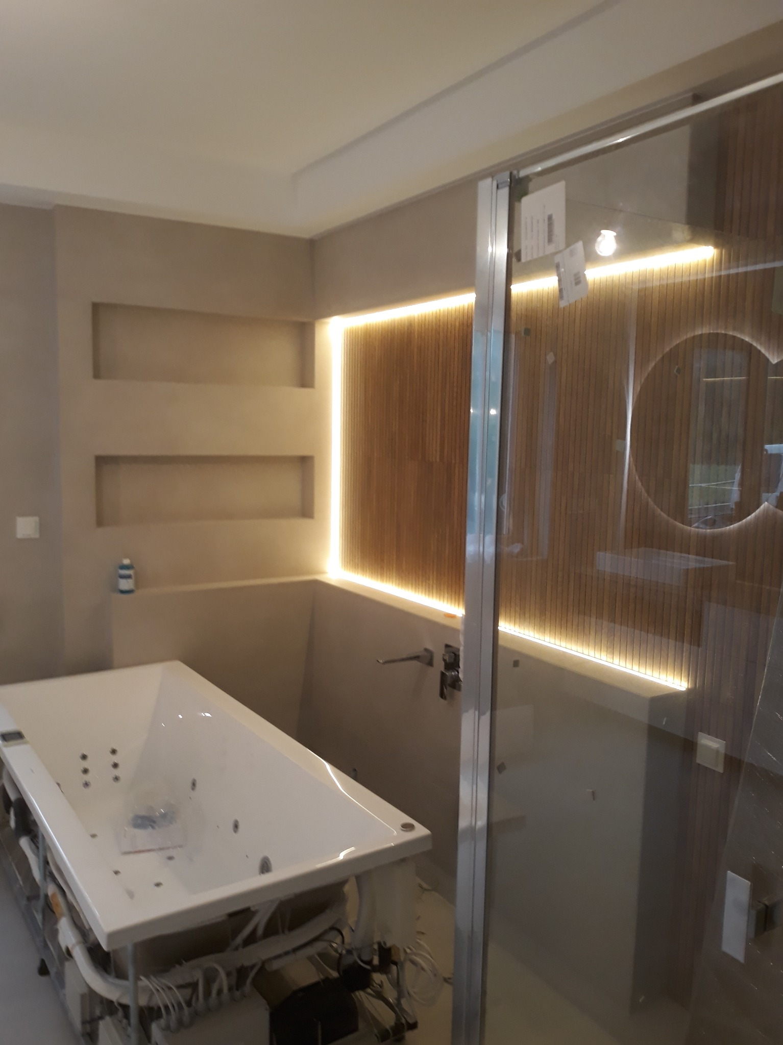 Nowoczesna łazienka w trakcie montażu: biała wanna z hydromasażem, szklana kabina prysznicowa z podświetlanymi panelami drewnianymi. Widoczne elementy instalacji.
