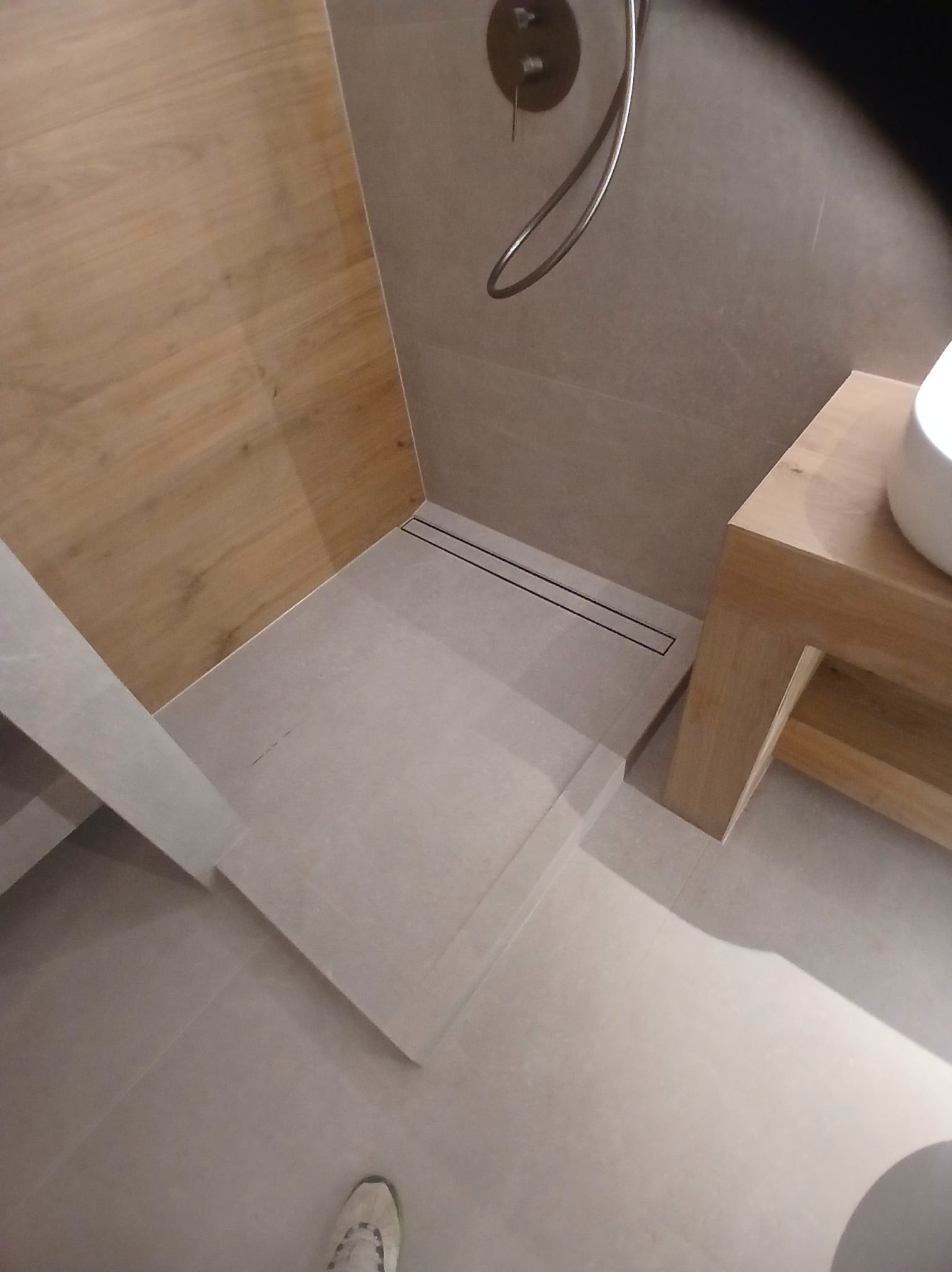 Nowoczesna kabina prysznicowa z odpływem liniowym, wykończona płytkami w odcieniach szarości i drewna. Minimalistyczny design z drewnianą umywalką i chromowaną armaturą.