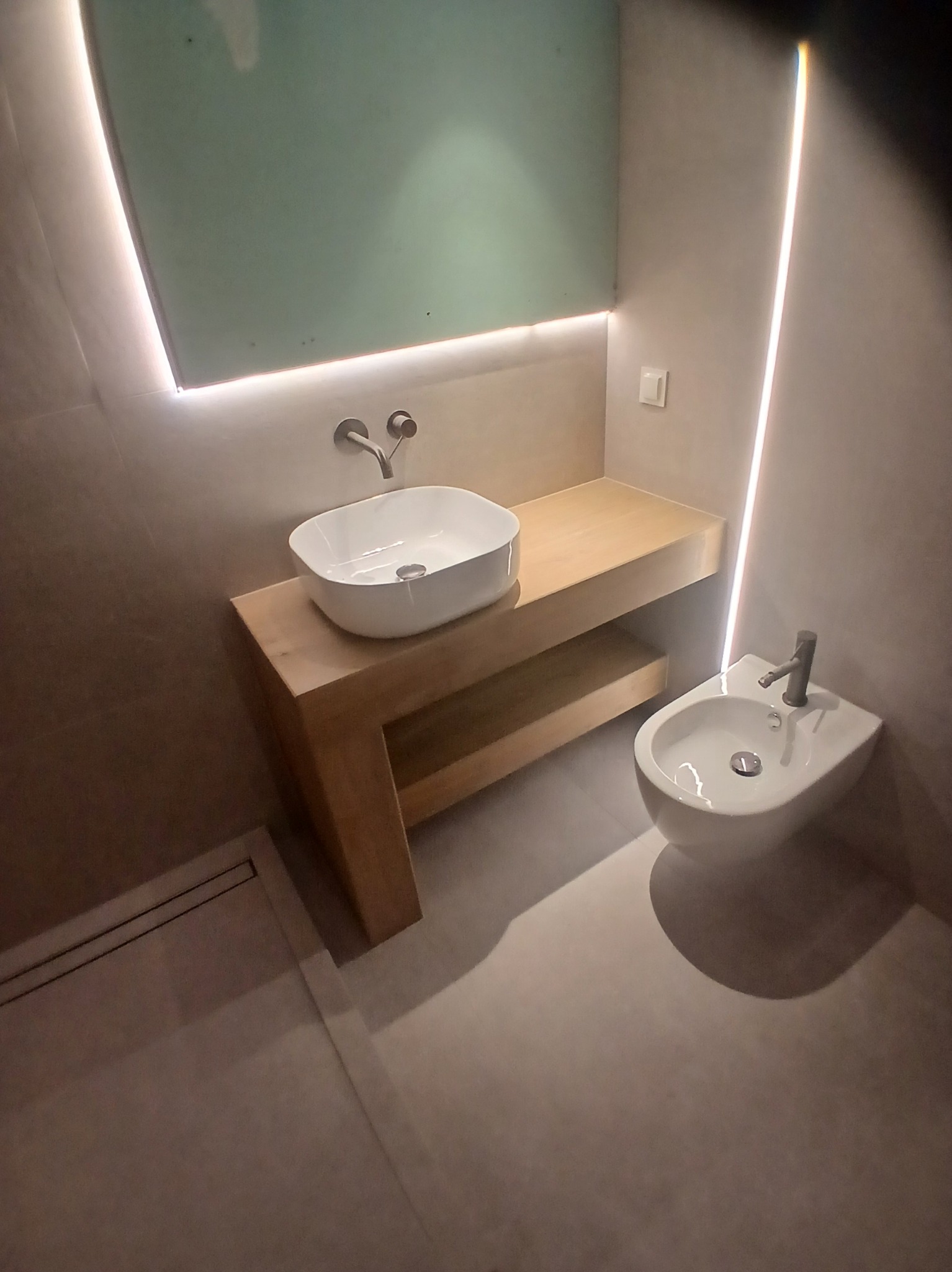 Nowoczesna łazienka z umywalką nablatową na drewnianym blacie i bidetem, oświetlona listwami LED. Minimalistyczny design, jasne kolory, eleganckie wykończenie.