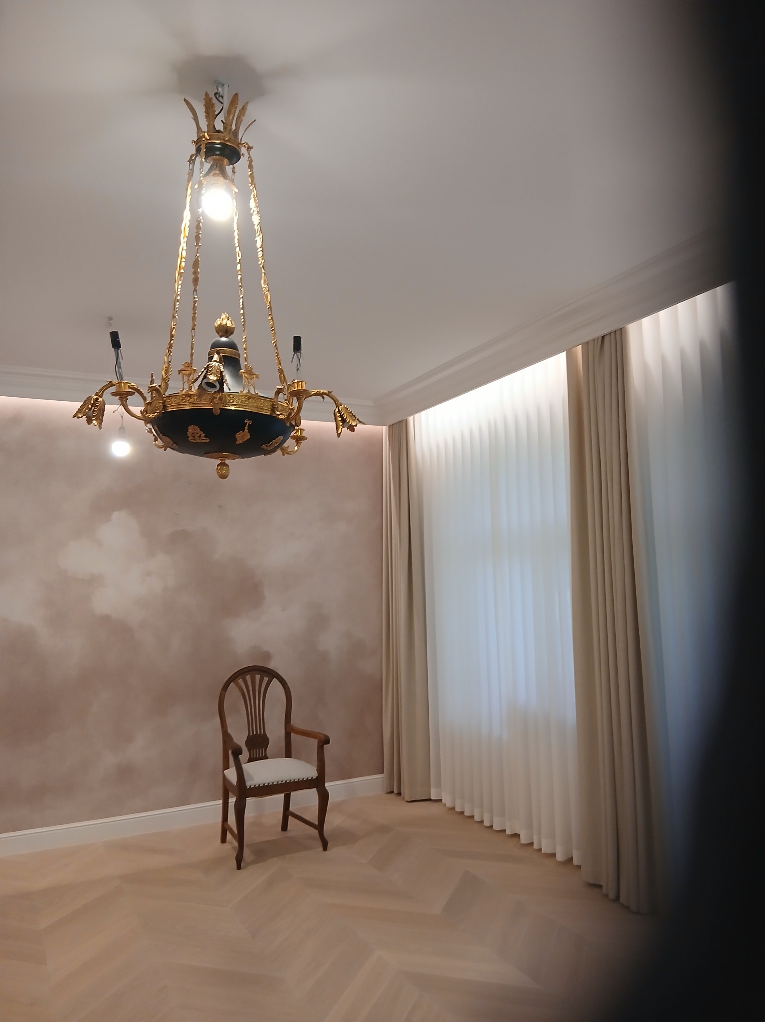 Eleganckie wnętrze z parkietem w jodełkę, krzesłem i ozdobnym żyrandolem. Ściana z efektem chmur i jasne zasłony w tle. Całość w stonowanej kolorystyce.