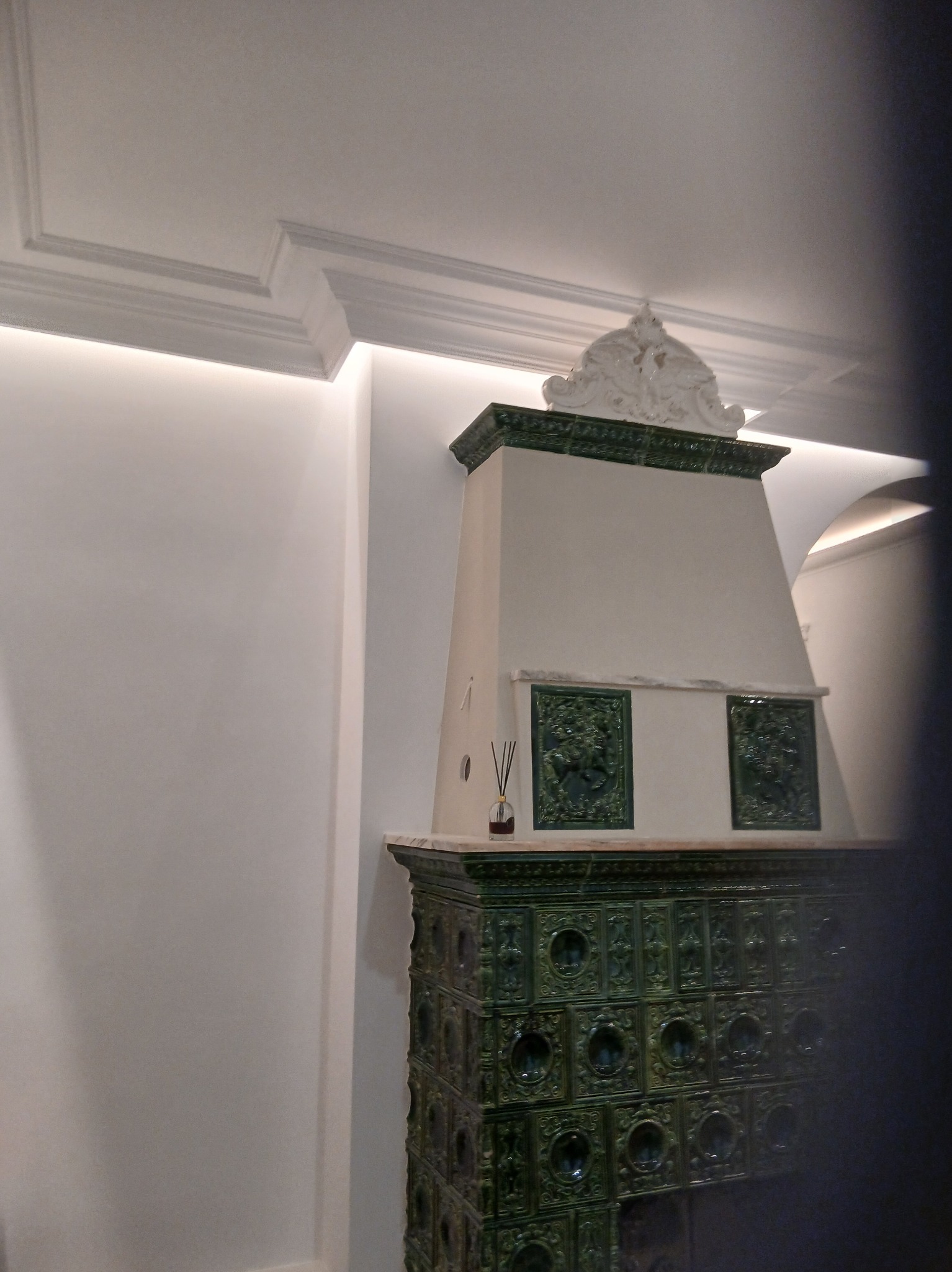 Stylowy, odnowiony piec kaflowy w odcieniach zieleni, z dekoracyjnymi kaflami i białym wykończeniem, w jasnym wnętrzu z subtelnym oświetleniem.