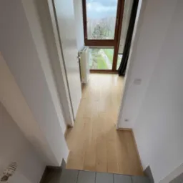 Jasne wnętrze domu z drewnianą podłogą i dużym oknem z widokiem na zieleń. Widoczny grzejnik i fragment schodów z szarymi płytkami.