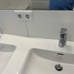 Natalia Dacyna - Biała, podwójna umywalka z chromowanymi bateriami i gniazdkami elektrycznymi w tle. Widoczne odbicie osoby z rolką papieru.