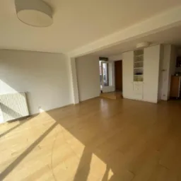 Jasne, puste wnętrze z drewnianą podłogą, białymi ścianami i szafą w zabudowie. Promienie słoneczne wpadają przez okno, tworząc geometryczne cienie na podłodze.