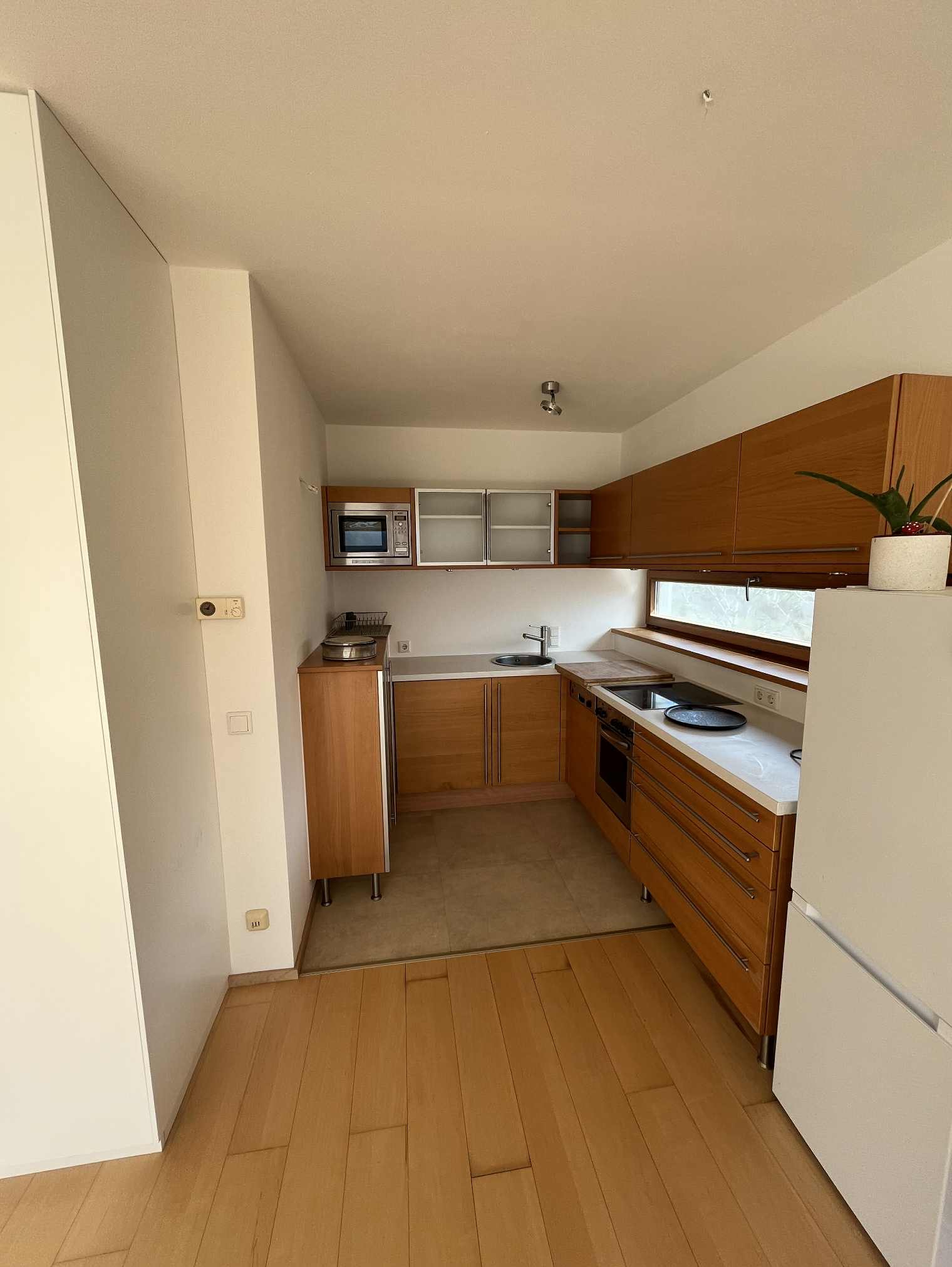 Jasna kuchnia z drewnianymi szafkami i białym blatem. Widoczne sprzęty AGD i roślina w doniczce. Parkiet na podłodze, minimalistyczny styl.