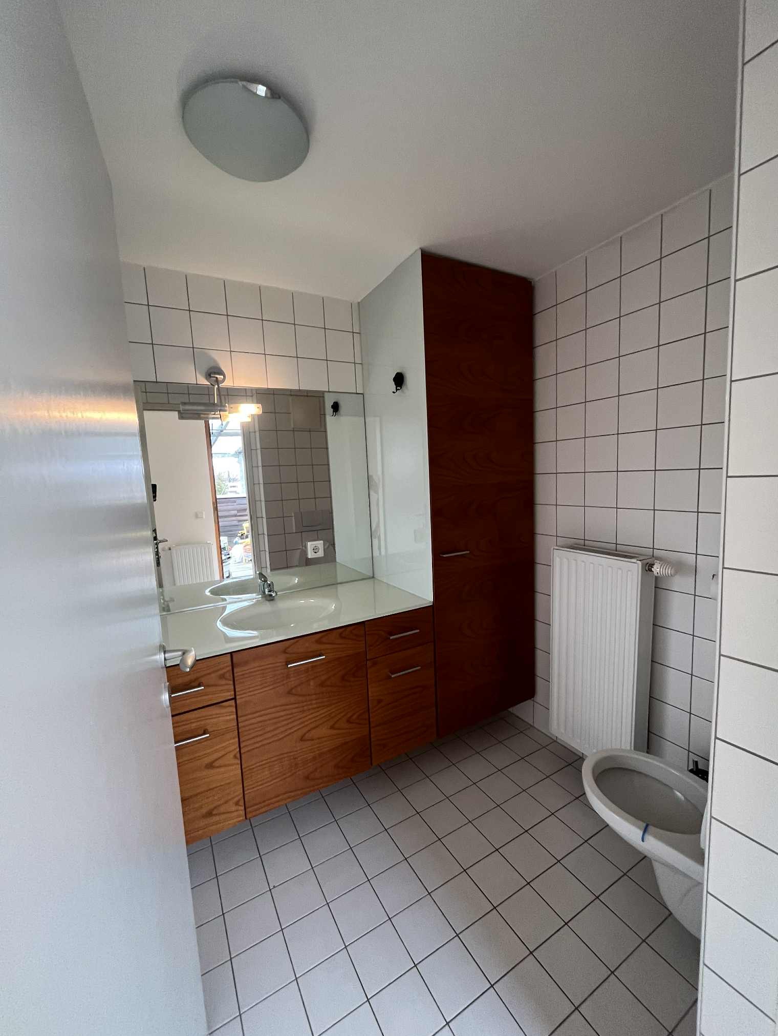 Wykończona łazienka z drewnianą szafką pod umywalką i białymi kwadratowymi płytkami na ścianach i podłodze. Widoczny również grzejnik i toaleta.