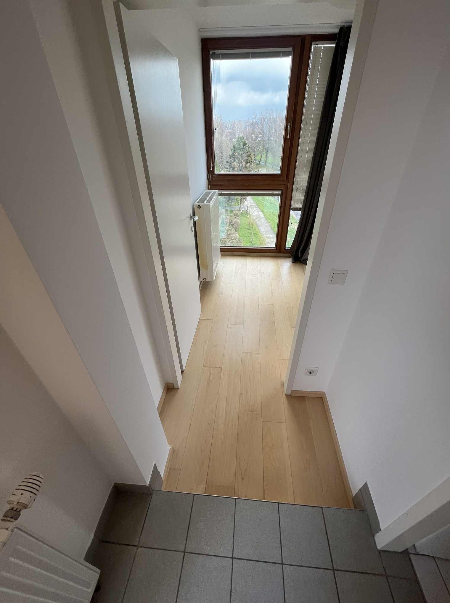 Jasne wnętrze domu z drewnianą podłogą i dużym oknem z widokiem na zieleń. Widoczny grzejnik i fragment schodów z szarymi płytkami.