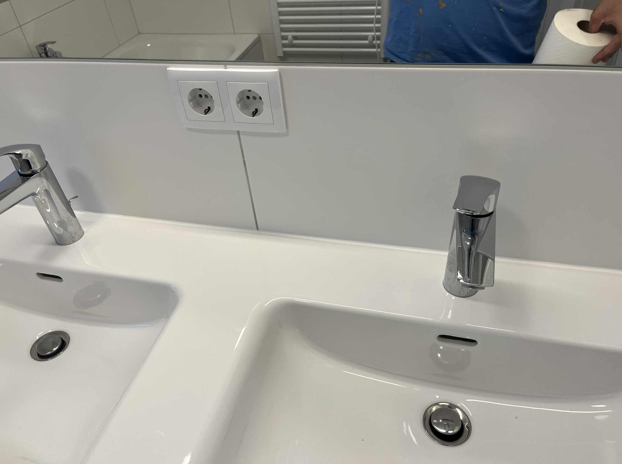 Biała, podwójna umywalka z chromowanymi bateriami i gniazdkami elektrycznymi w tle. Widoczne odbicie osoby z rolką papieru.