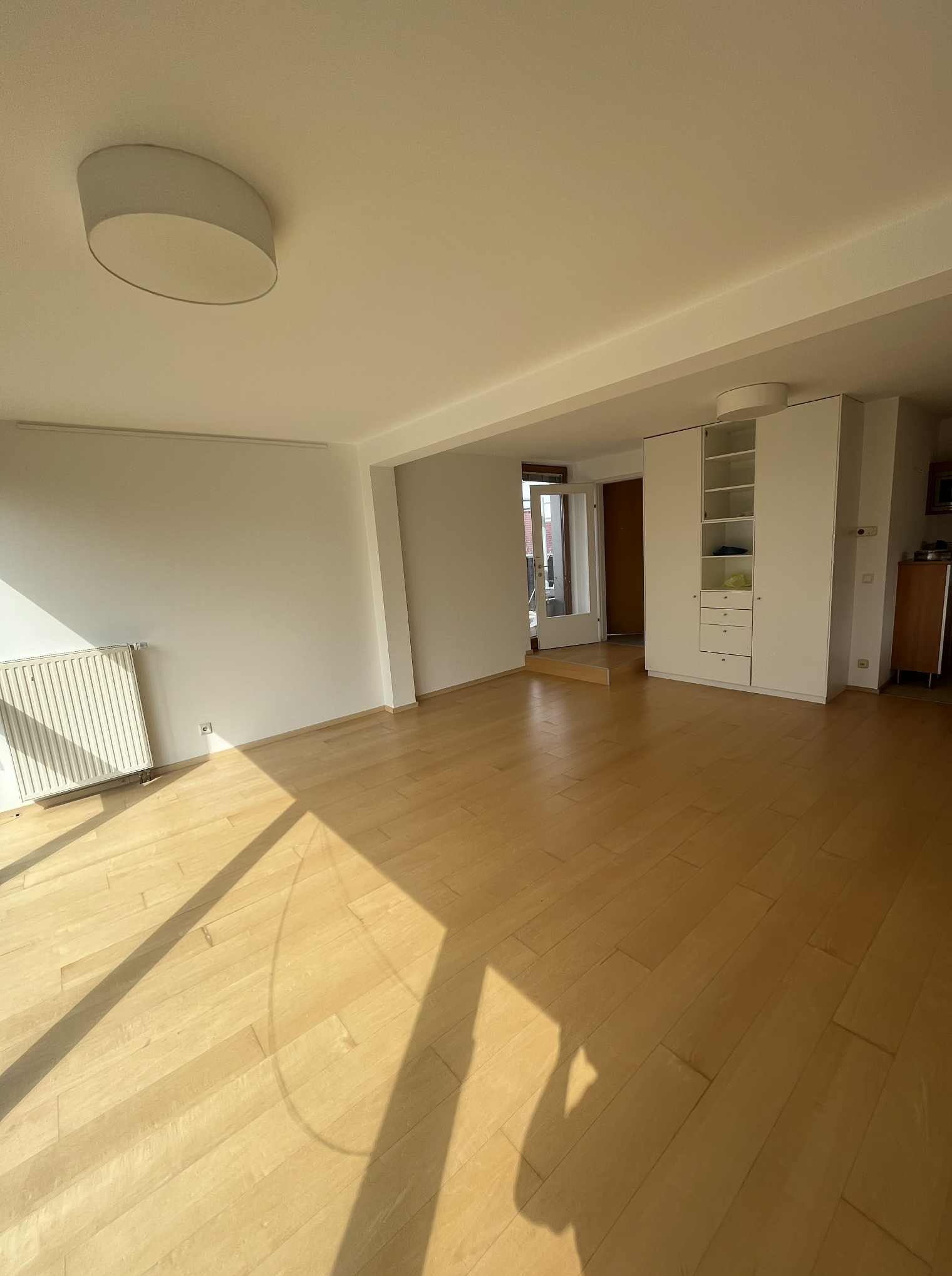 Jasne, puste wnętrze z drewnianą podłogą, białymi ścianami i szafą w zabudowie. Promienie słoneczne wpadają przez okno, tworząc geometryczne cienie na podłodze.