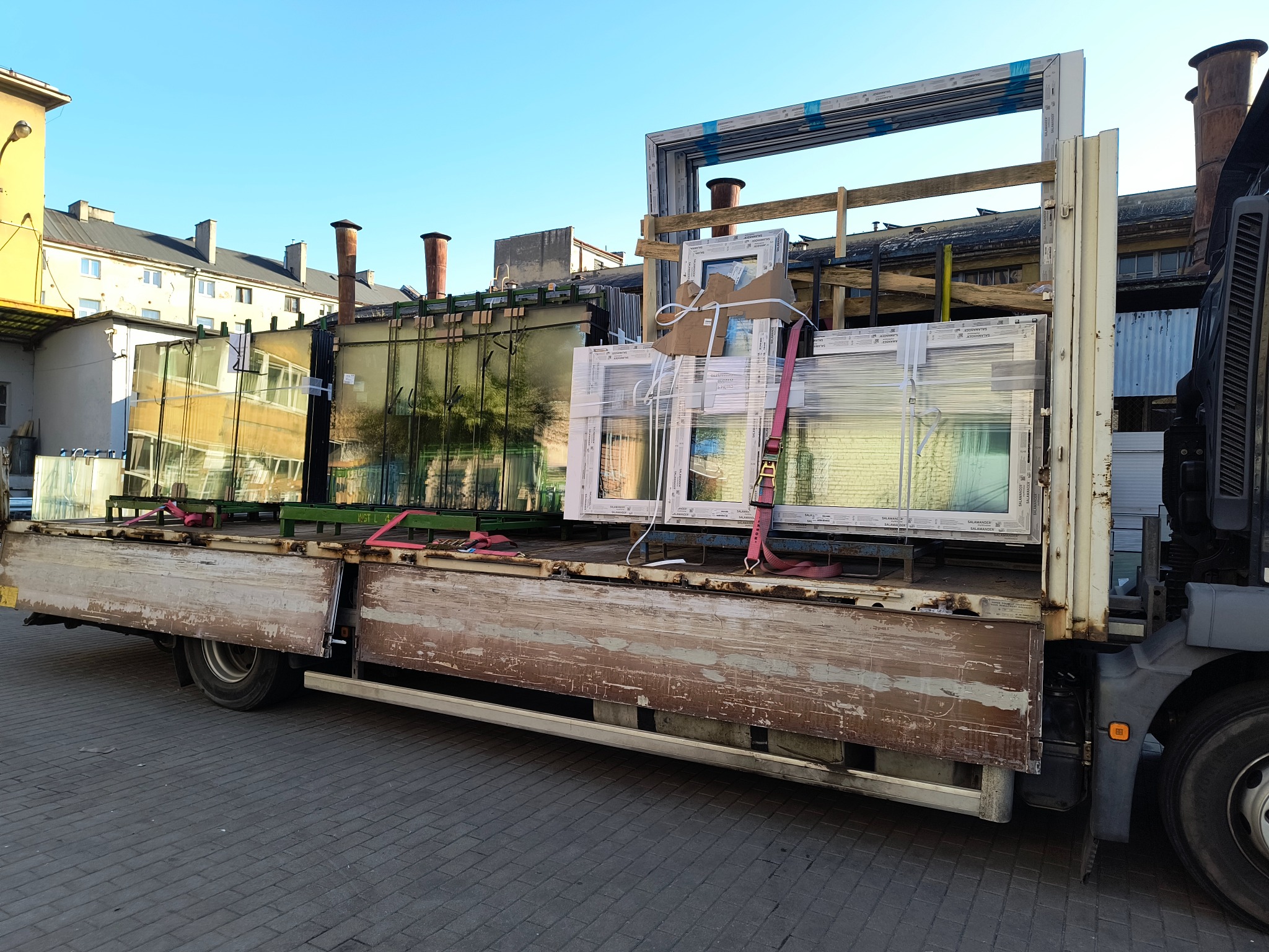 Transport okien Salamander na ciężarówce z otwartą platformą, zabezpieczonych pasami. Widoczne budynki w tle i niebo.