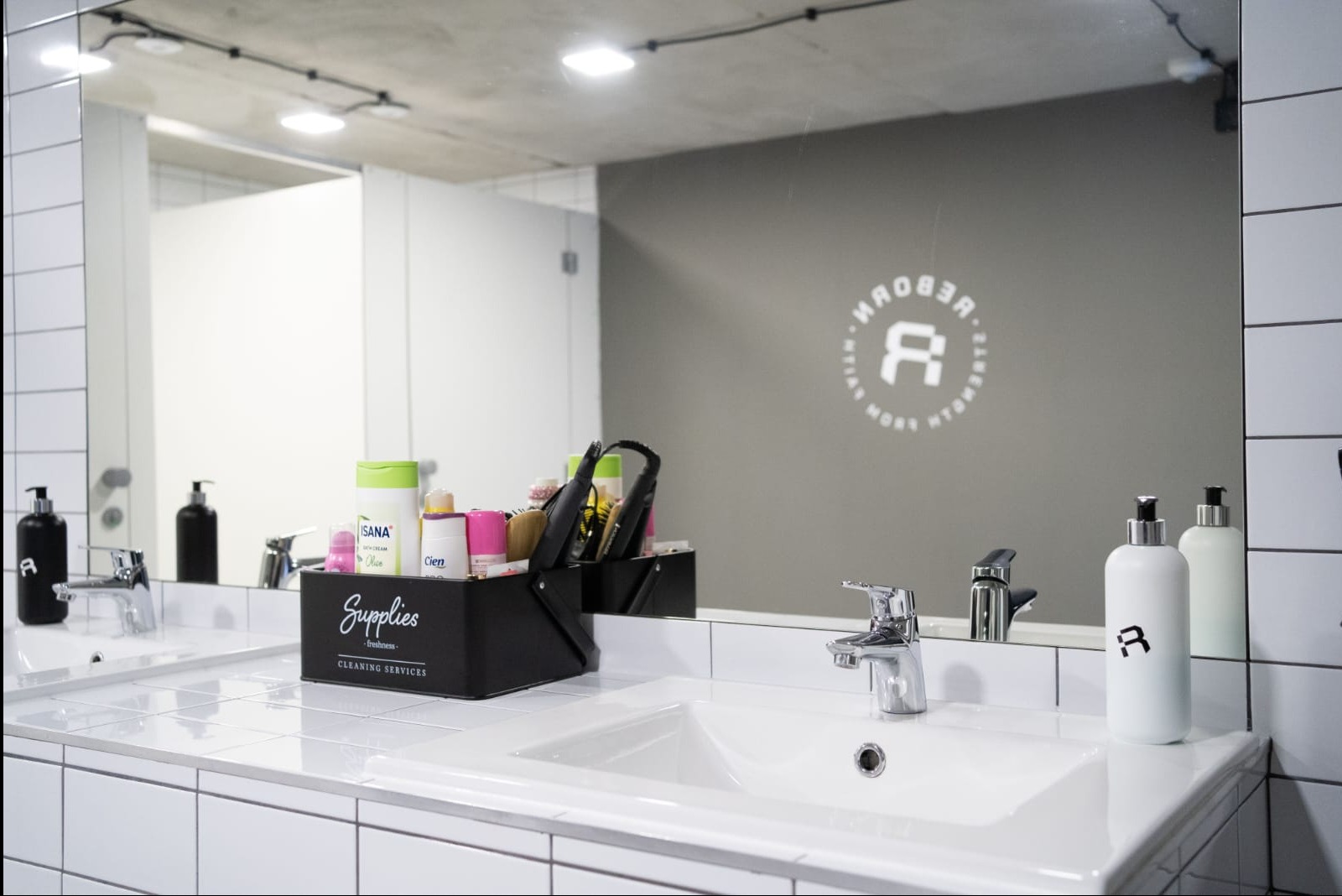 Nowoczesna łazienka z białymi płytkami i akcesoriami treningowymi w organizerze, odbijającymi się w dużym lustrze z logo siłowni.