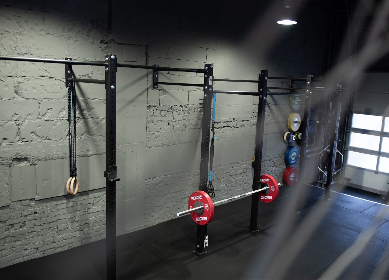 Nowoczesna siłownia z czarnymi stojakami do ćwiczeń, obciążeniami i gimnastycznymi kółkami na tle szarej, ceglanej ściany. Oświetlenie sufitowe, gumowa podłoga.