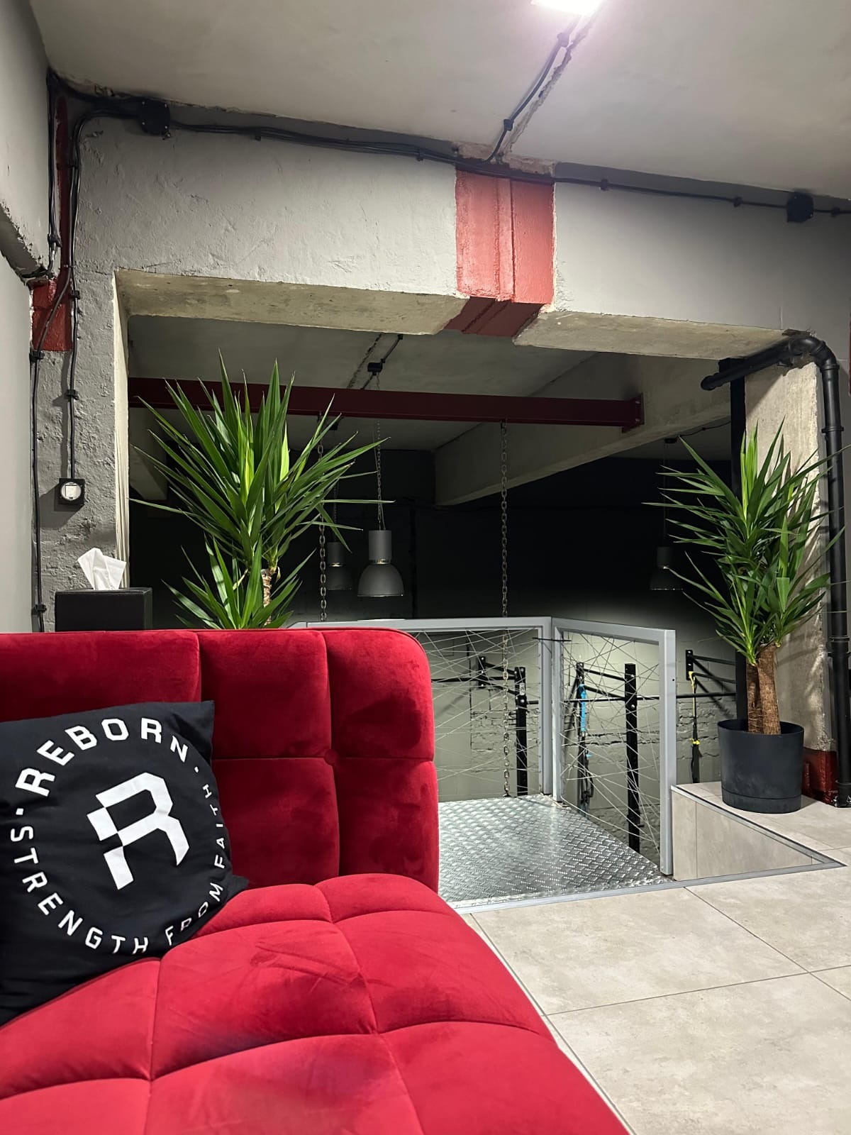 Czerwona sofa z poduszką 'Reborn' w surowym wnętrzu siłowni z roślinami, widok na salę treningową z metalowymi konstrukcjami i łańcuchami.