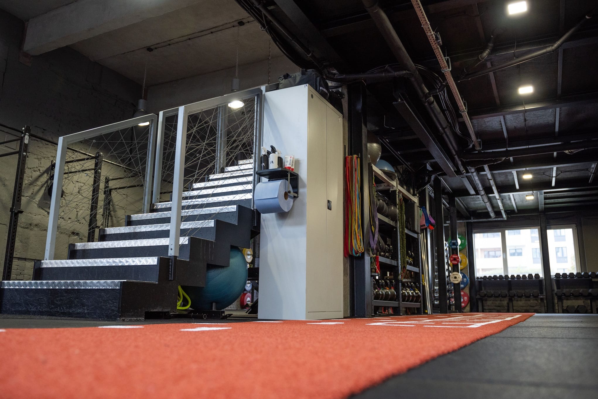 Widok na nowoczesną siłownię: schody z metalowymi stopniami, regały z obciążeniami, maty podłogowe i okno. Widoczne akcesoria do ćwiczeń i dbałość o czystość.