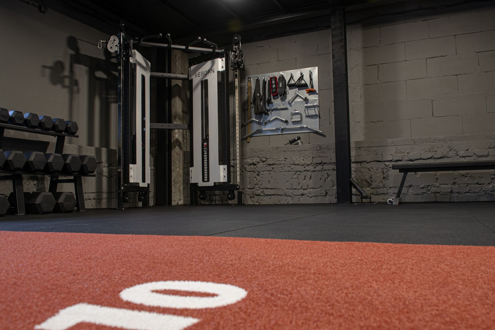 Nowoczesna siłownia z profesjonalnym sprzętem: maszyna do ćwiczeń, hantle na stojaku i akcesoria na ścianie z cegły. Czerwona bieżnia z białym napisem na podłodze.