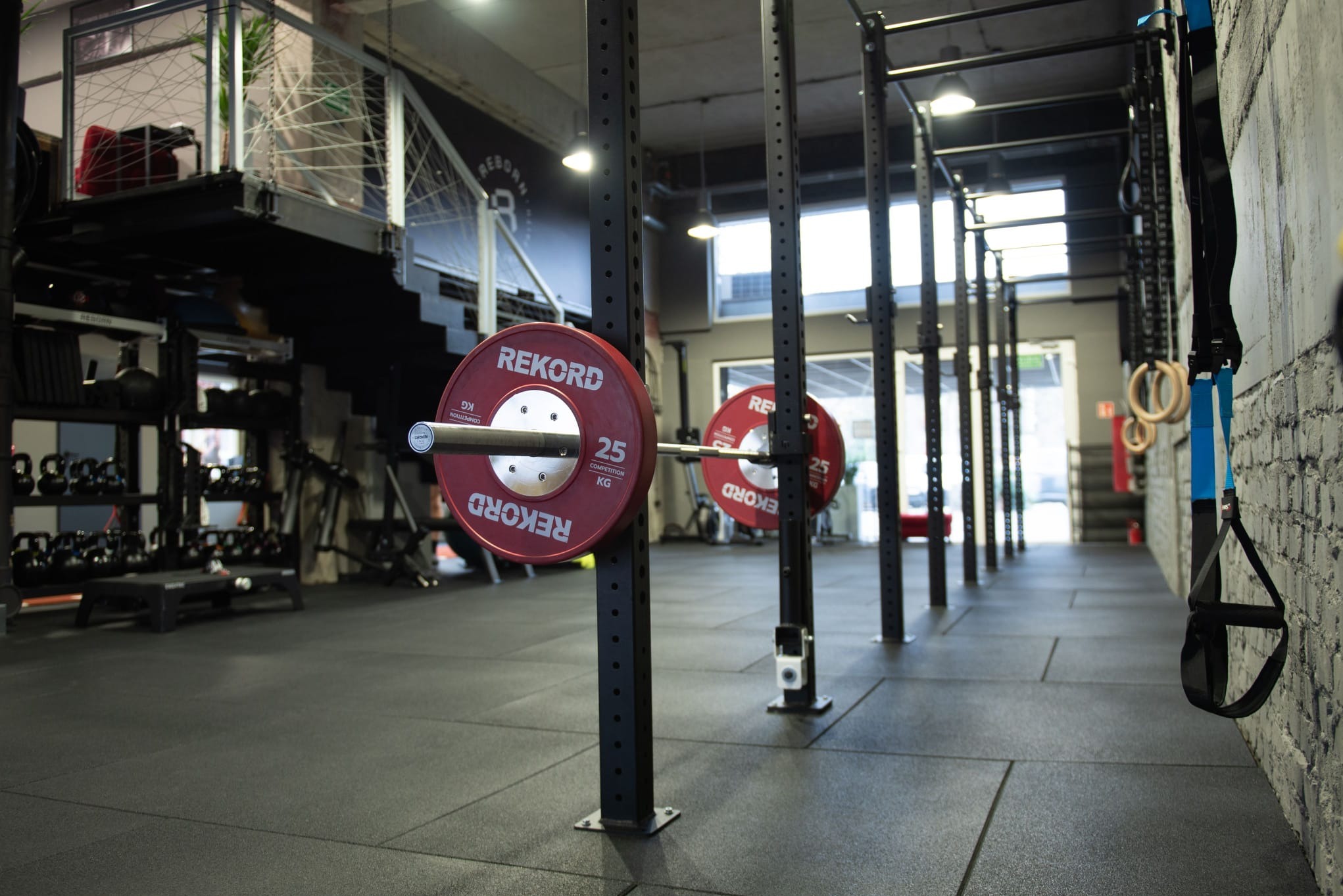 Wnętrze siłowni z ciężarami Rekord 25 kg na stojakach, widok na korytarz ze sprzętem do ćwiczeń i drabinkami. Nowoczesny design, profesjonalny sprzęt.