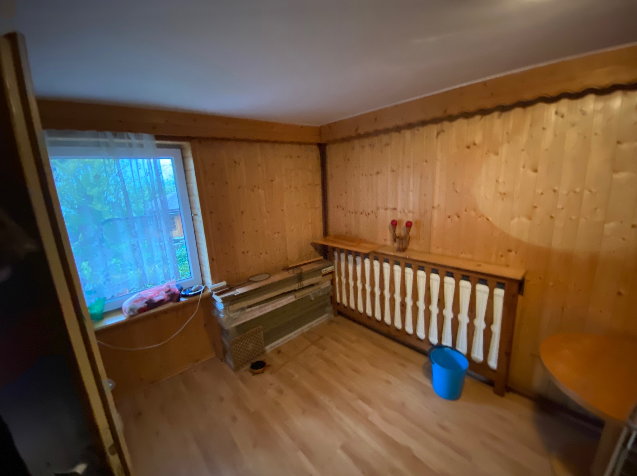 Pokój wyłożony boazerią z oknem, przy którym leżą spakowane kartony. Przy grzejniku stoi niebieskie wiadro, co sugeruje przygotowania do opróżniania.