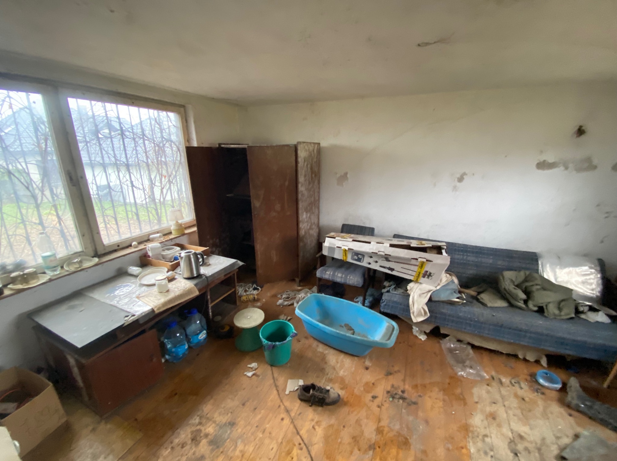 Wnętrze zaniedbanego pokoju z meblami, brudną podłogą i oknem. Widoczne ślady użytkowania, stare meble i przedmioty codziennego użytku. Generalny nieporządek.