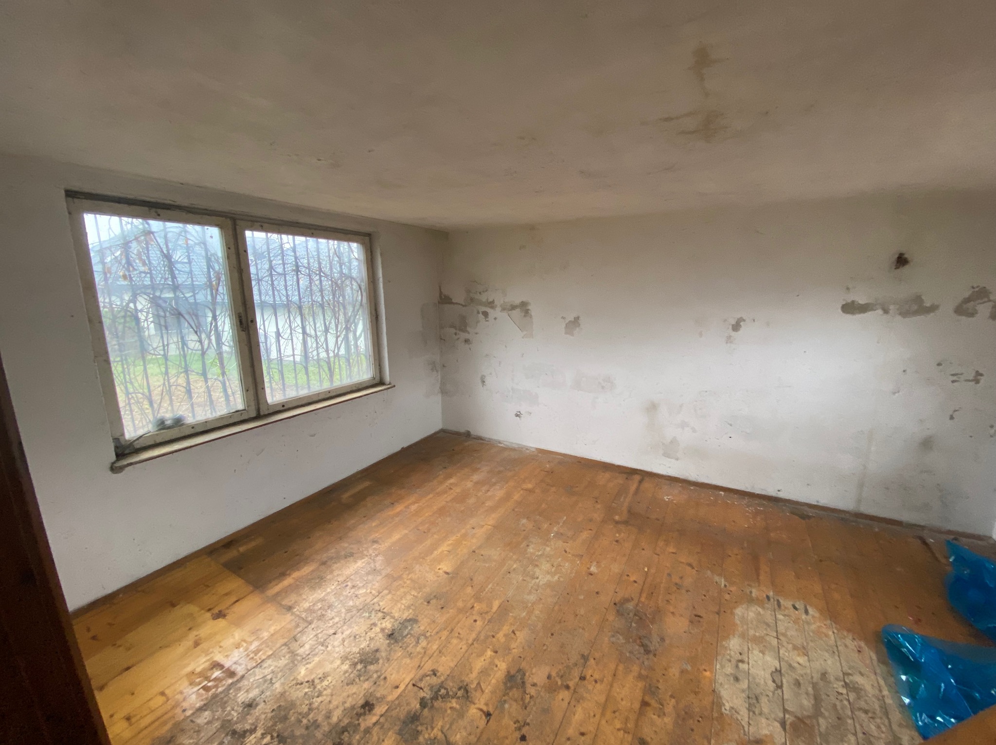 Wnętrze zaniedbanego pokoju z drewnianą podłogą, widoczne ślady wilgoci na ścianach i podłodze, okno z kratą i worki na śmieci w rogu.