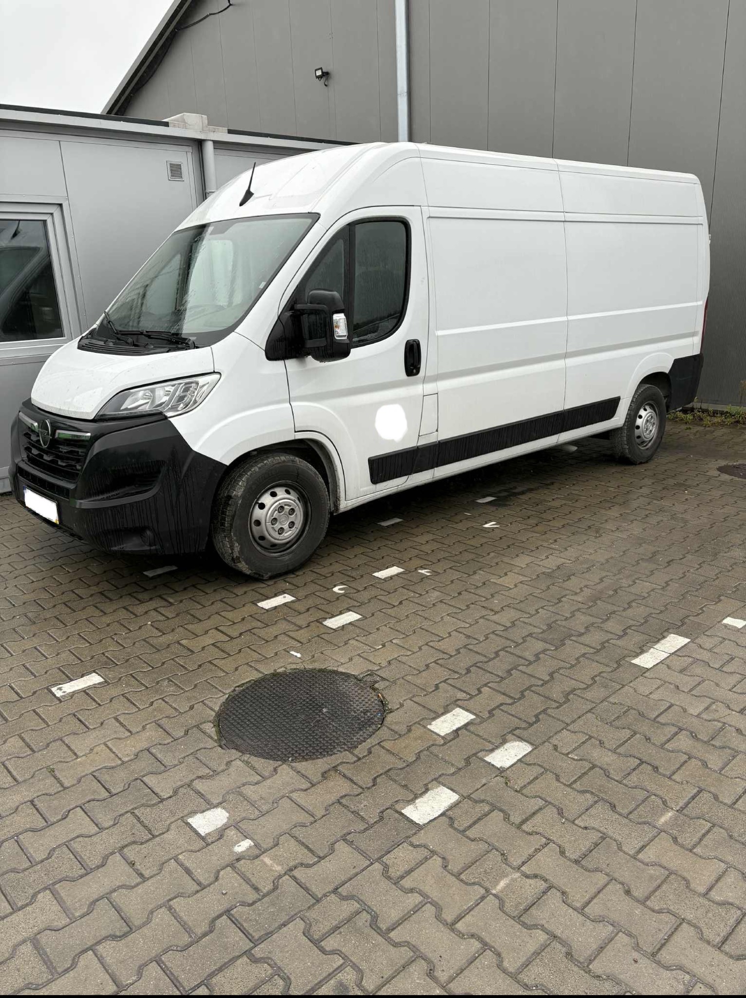 Biały bus dostawczy zaparkowany na kostce brukowej. Widoczny z boku, na tle szarego budynku. Kostka z białymi liniami wyznaczającymi miejsca parkingowe.