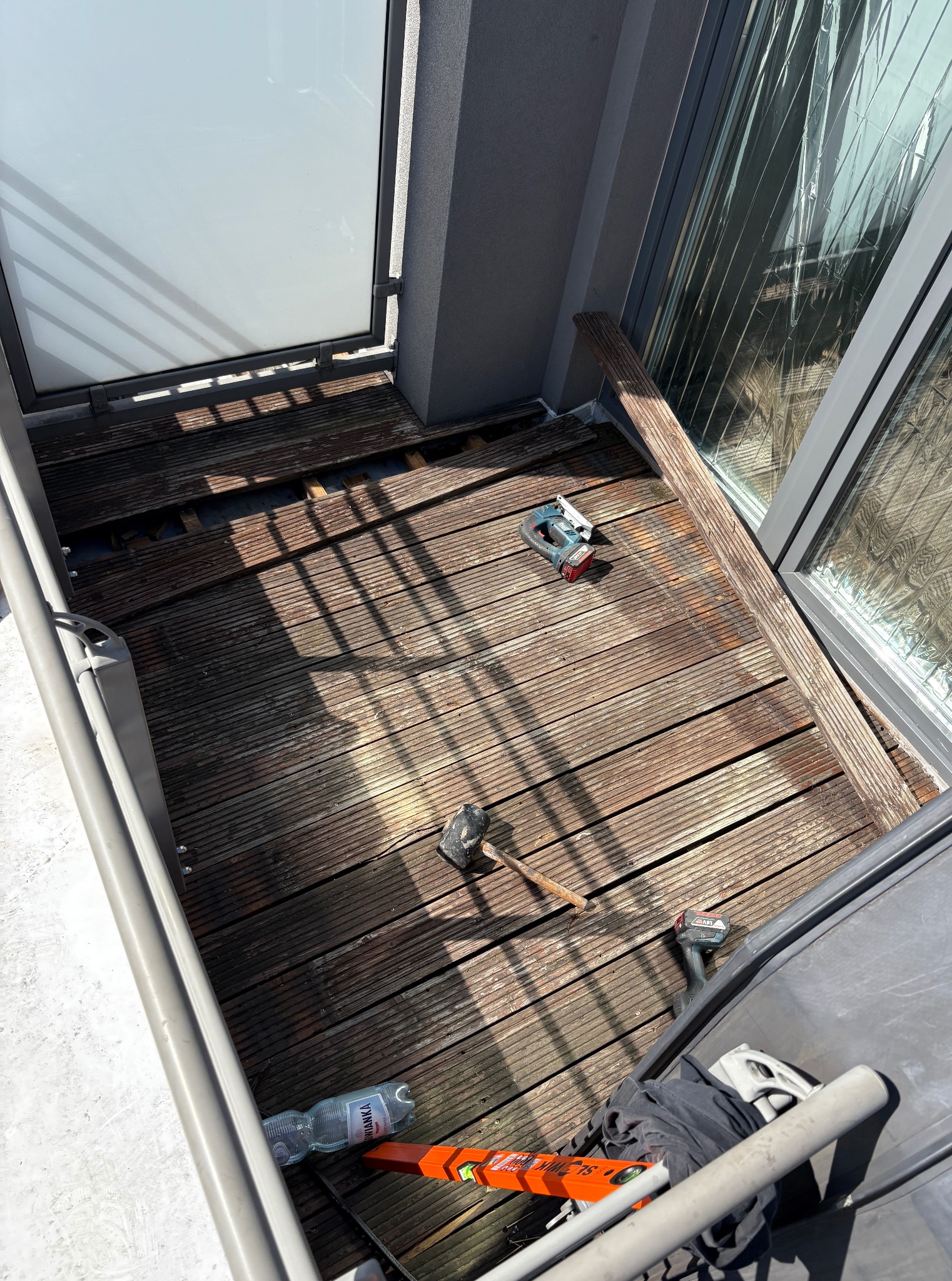 Demontaż starego tarasu drewnianego na balkonie z widocznymi narzędziami: wyrzynarka, poziomica, młotek. Częściowo usunięte deski tarasowe. Prace w toku.