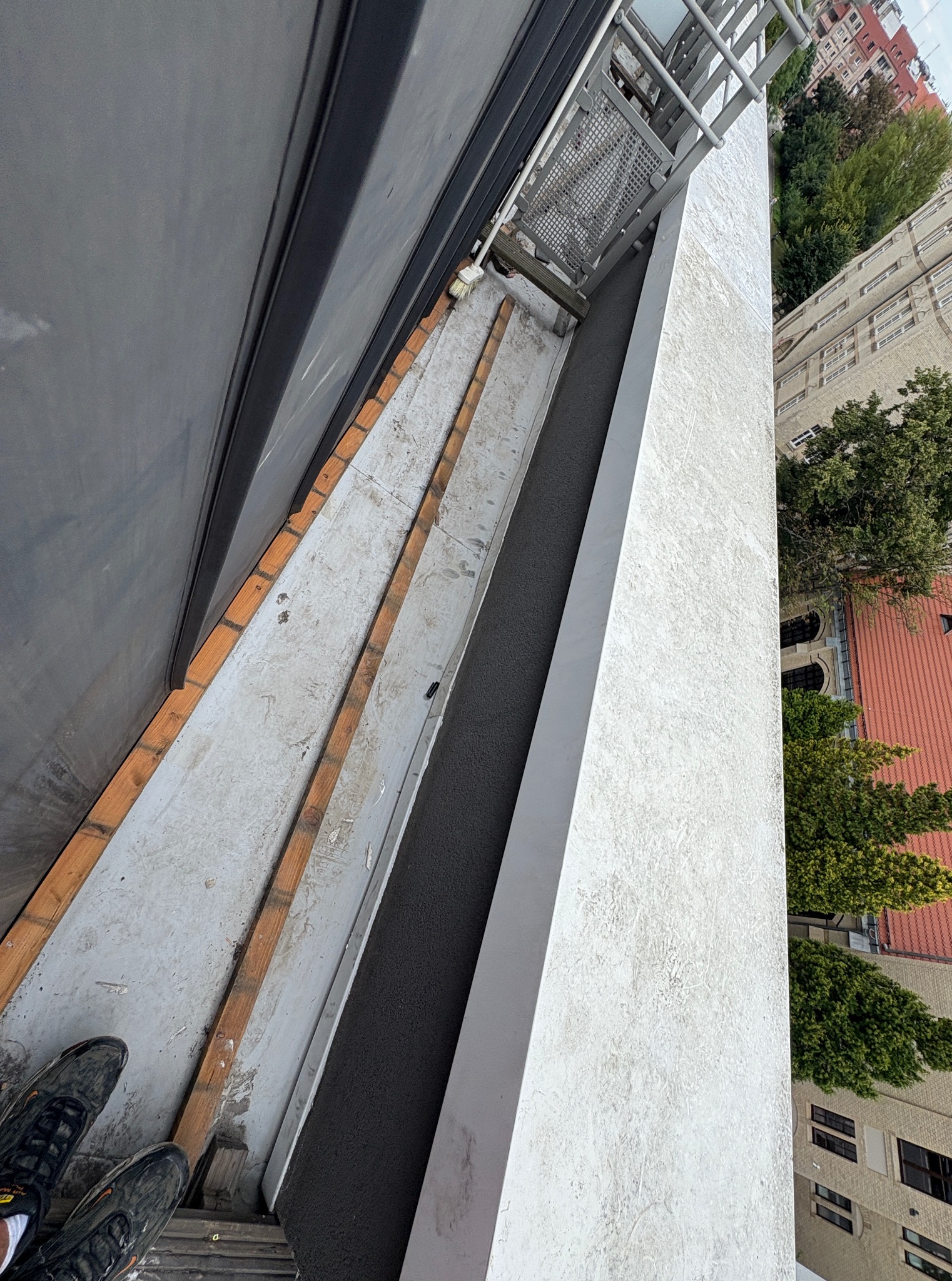 Widok z góry na balkon w trakcie prac. Widoczne elementy konstrukcyjne, fragment ściany z fakturą i fragment butów osoby fotografującej. W tle budynki i zieleń.