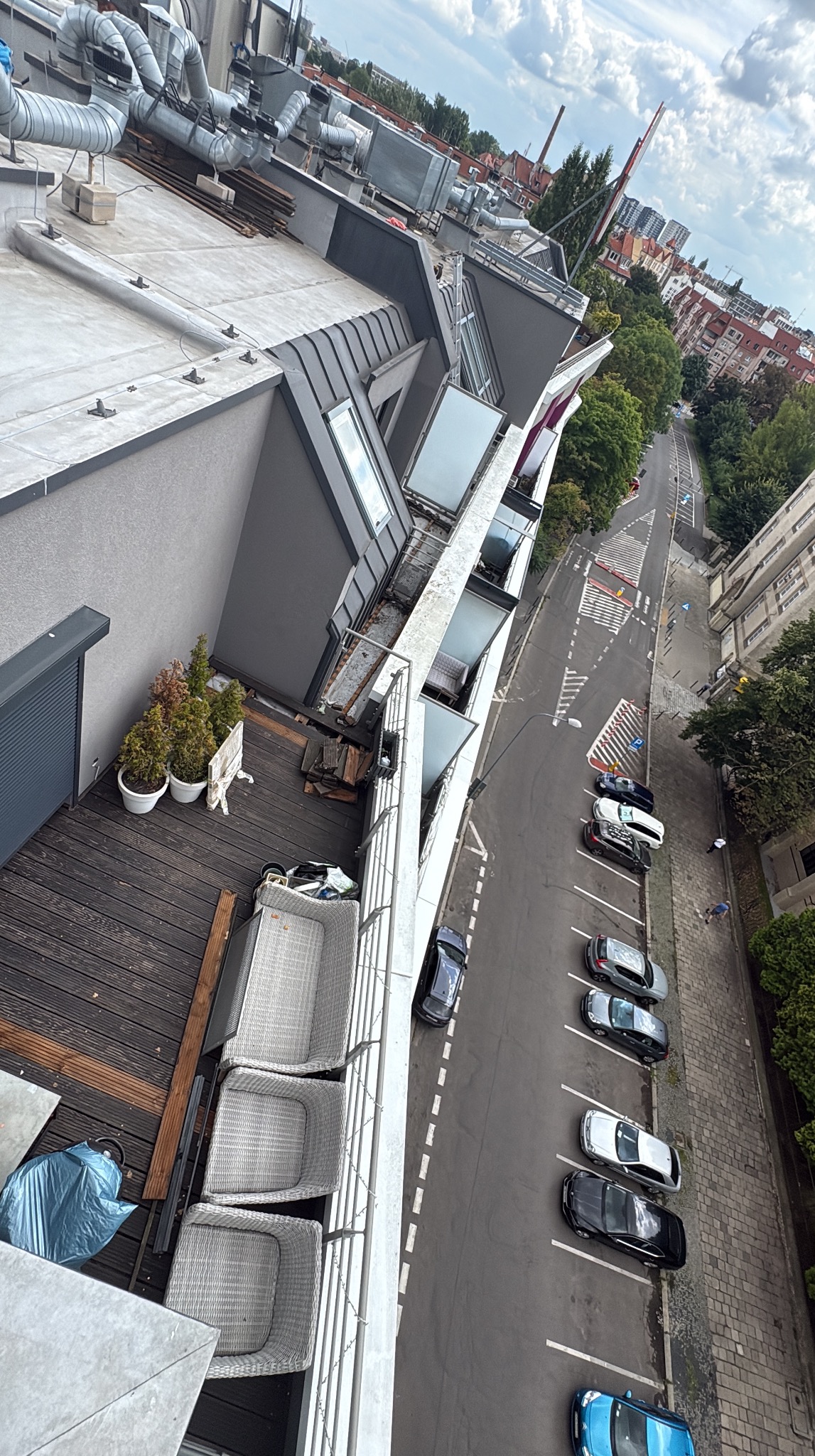 Widok z góry na balkon z drewnianą podłogą i rattanowymi meblami, zza balustrady widoczna ulica z zaparkowanymi samochodami i budynki w tle.