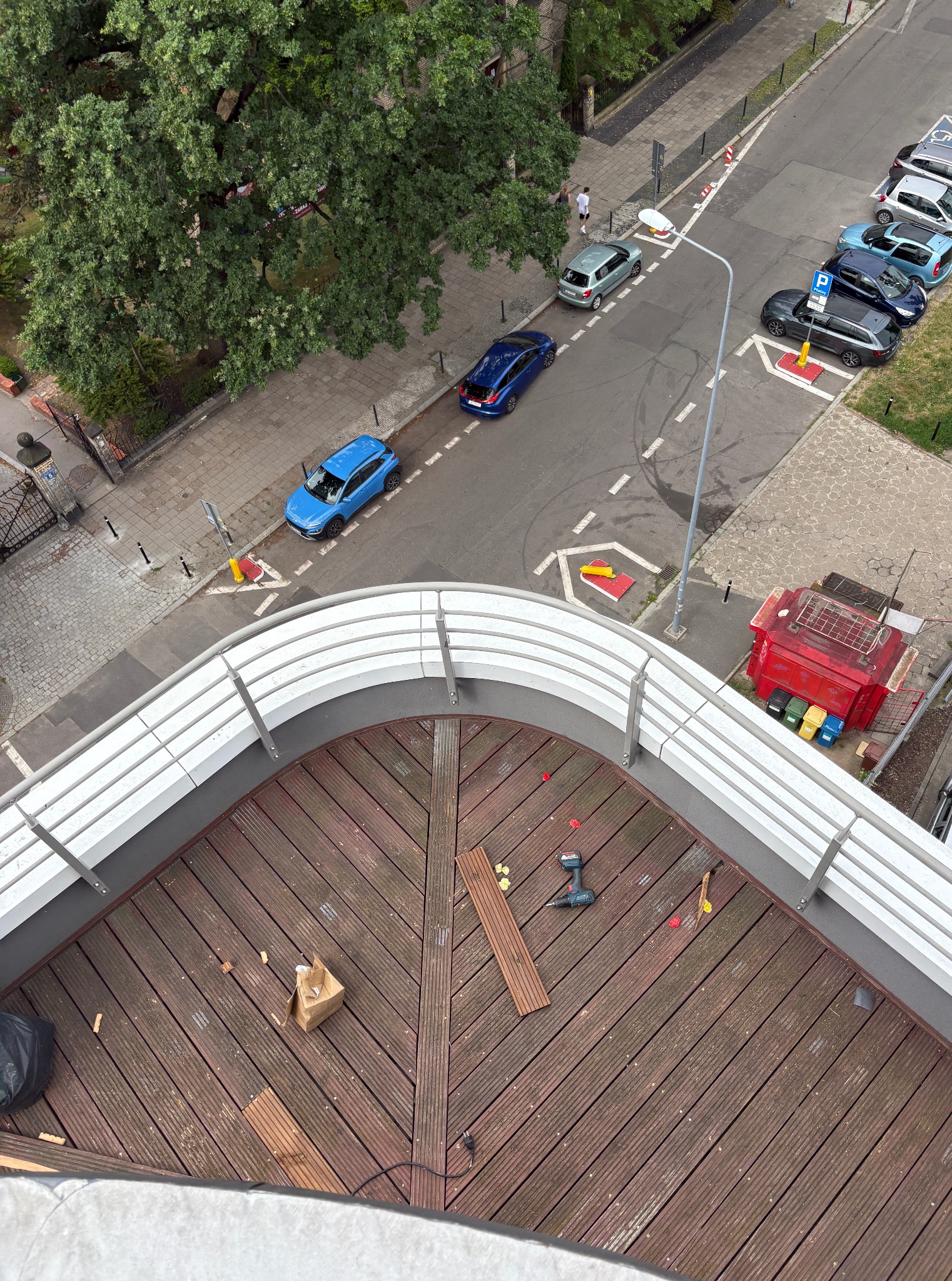 Widok z góry na taras z desek kompozytowych w trakcie montażu, z narzędziami i elementami mocującymi. W tle ulica z zaparkowanymi autami i drzewa.