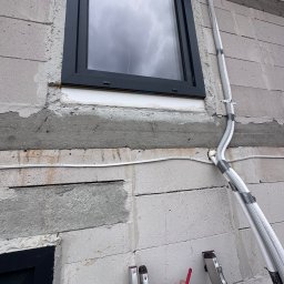 Energo-Tech Bednarczyk - Montaż rur gazowych przy oknie w surowym budynku z betonu komórkowego. Rury zabezpieczone taśmą, przymocowane uchwytami. Widoczna drabina i dłoń z markerem.