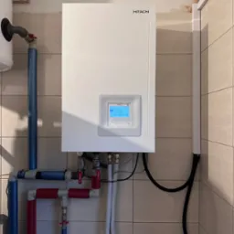 Biała pompa ciepła Hitachi zamontowana na ścianie z beżowych płytek, widoczne rury w kolorze niebieskim i czerwonym. Na wyświetlaczu pompy widoczna temperatura.