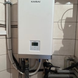 Energo-Tech Bednarczyk - Biała pompa ciepła Kaisai zamontowana na ścianie z beżowych płytek, widoczne rury i kable. Kompaktowy model z wyświetlaczem i przyciskami.