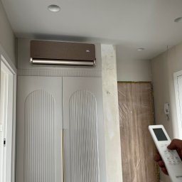 Energo-Tech Bednarczyk - Brązowy klimatyzator na ścianie nad białą szafą z dekoracyjnymi frezami. W prawej części kadru dłoń trzyma pilot. Widoczne ślady prac wykończeniowych.