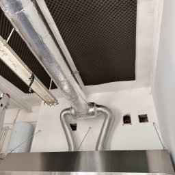 Energo-Tech Bednarczyk - Srebrny przewód wentylacyjny z izolacją termiczną, podłączony do okapu kuchennego w lokalu gastronomicznym. Na suficie panele akustyczne.