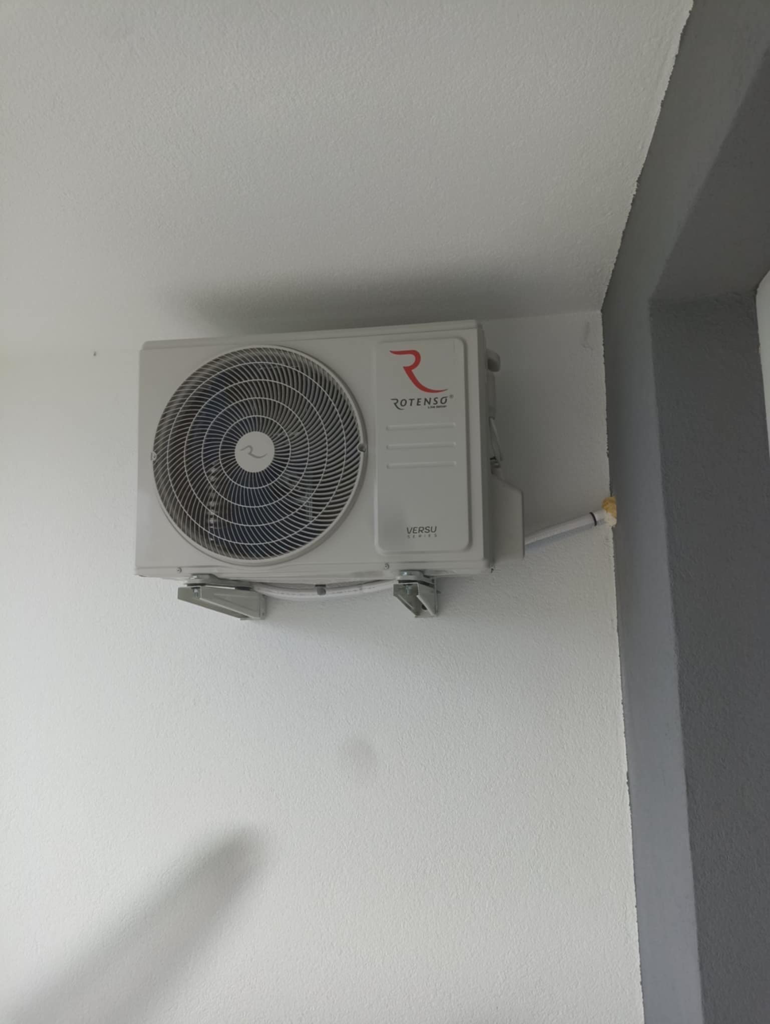 Klimatyzator Rotenso Versu Series zamontowany na białej ścianie, widoczne rurki instalacyjne. Kąt ujęcia z dołu uwydatnia mocowanie urządzenia.