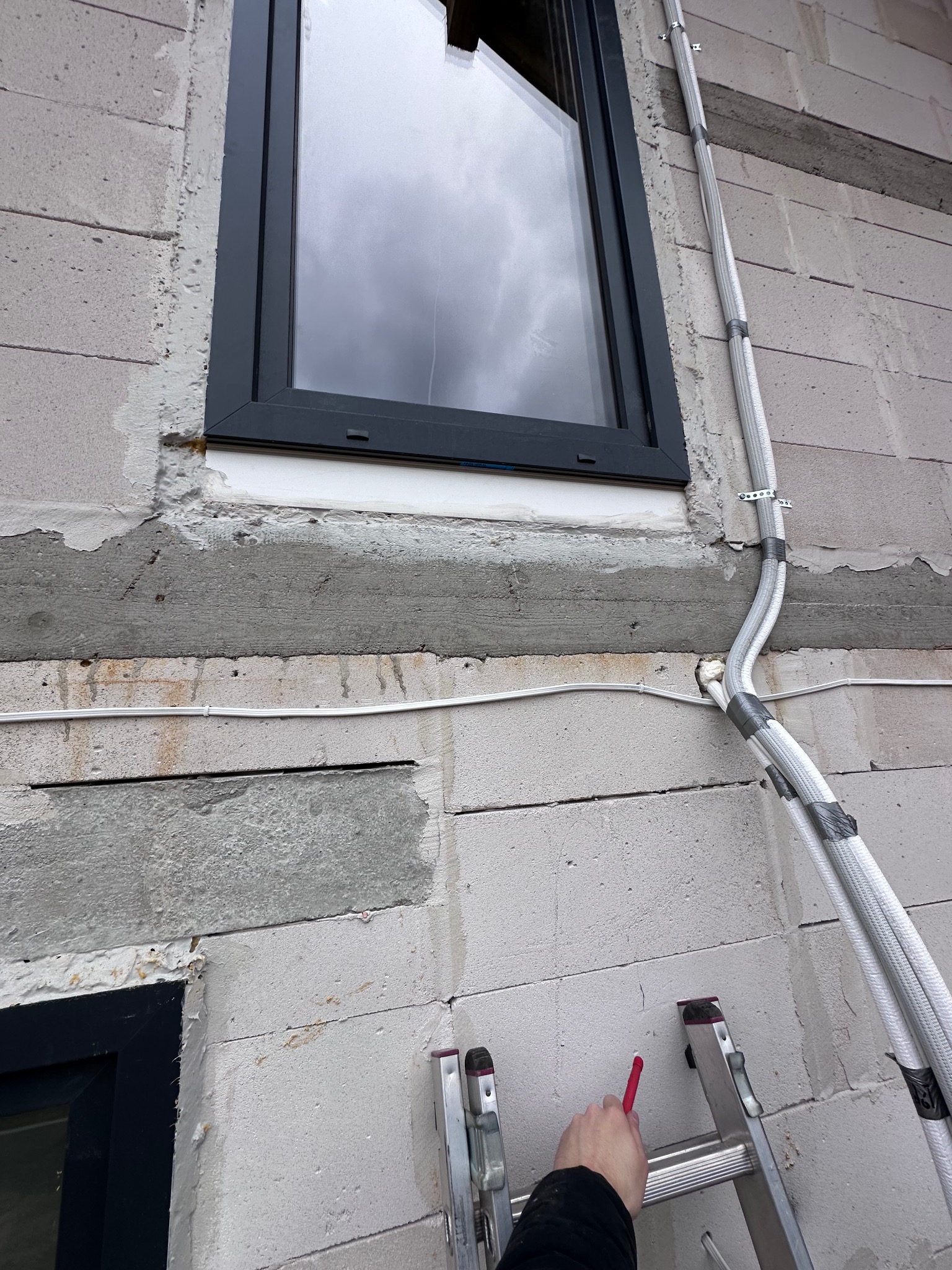Montaż rur gazowych przy oknie w surowym budynku z betonu komórkowego. Rury zabezpieczone taśmą, przymocowane uchwytami. Widoczna drabina i dłoń z markerem.