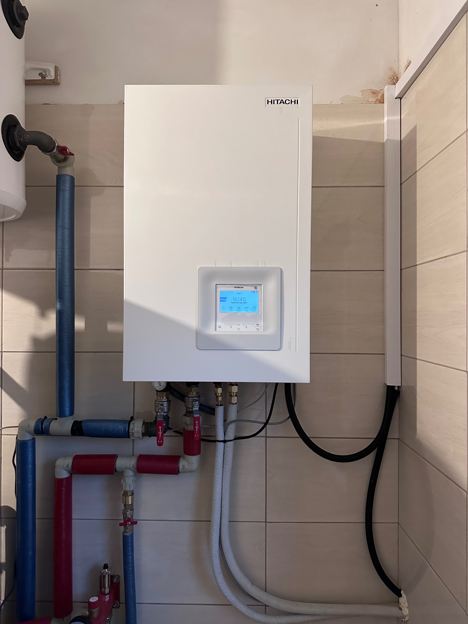 Biała pompa ciepła Hitachi zamontowana na ścianie z beżowych płytek, widoczne rury w kolorze niebieskim i czerwonym. Na wyświetlaczu pompy widoczna temperatura.