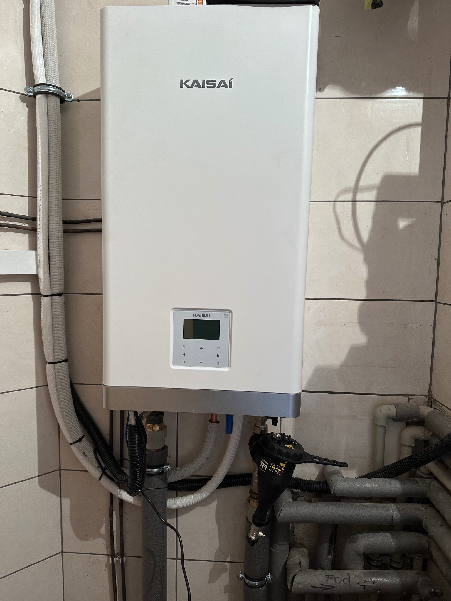 Biała pompa ciepła Kaisai zamontowana na ścianie z beżowych płytek, widoczne rury i kable. Kompaktowy model z wyświetlaczem i przyciskami.
