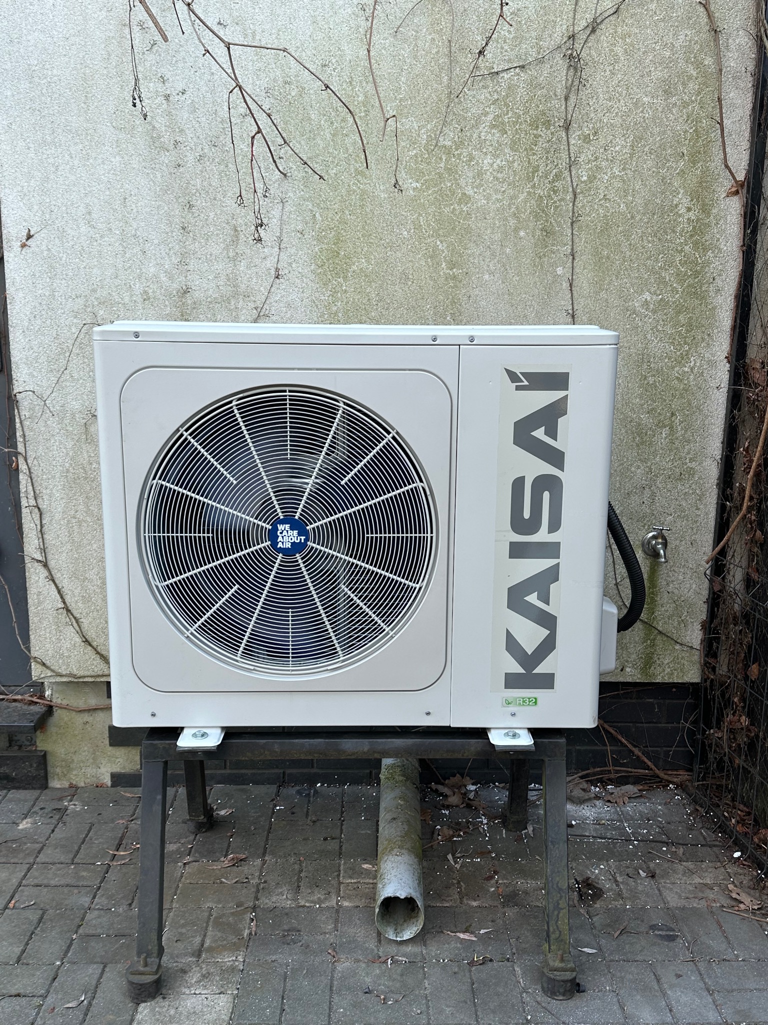 Biała jednostka zewnętrzna klimatyzacji Kaisai na metalowym stojaku przed ścianą z bluszczem. Widoczny kran i rura odpływowa.