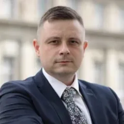 Portret mężczyzny w garniturze i krawacie w kwiaty na tle budynku. Krótkie włosy, poważny wyraz twarzy. Ujęcie od klatki piersiowej w górę. Delikatne oświetlenie.