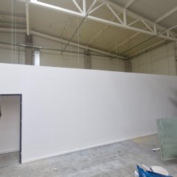 Maryan Mazan usługi budowlane - Biała ściana działowa w hali magazynowej z widocznym regałem, kartonami i materiałami budowlanymi. Oświetlenie sufitowe, betonowa posadzka.