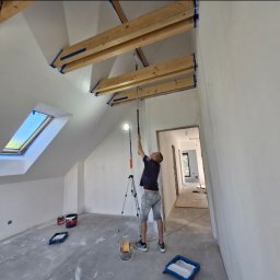 Maryan Mazan usługi budowlane - Malowanie sufitu z drewnianymi belkami w pomieszczeniu ze skosami i oknem dachowym. Widoczny malarz używający wałka na wysięgniku i lampy na statywie.
