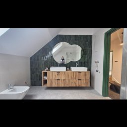 Maryan Mazan usługi budowlane - Nowoczesna łazienka na poddaszu z drewnianą szafką pod umywalki, zieloną glazurą i owalnym lustrem. W tle widoczne drzwi z zieloną ościeżnicą i bidet.