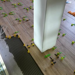 Maryan Mazan usługi budowlane - Układanie drewnopodobnych płytek podłogowych z użyciem klipsów poziomujących, widoczna warstwa kleju i fragment kolumny. Praca w toku, zbliżenie na podłogę.