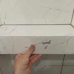 Maryan Mazan usługi budowlane - Układanie białych płytek imitujących marmur z brązowymi żyłkami. Widoczna ręka glazurnika przytrzymująca płytkę. Napis na płytce.