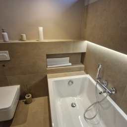 Maryan Mazan usługi budowlane - Nowoczesna łazienka w beżowych tonacjach: wanna, toaleta, półki z oświetleniem LED. Minimalistyczny design, ciepłe światło, płytki imitujące kamień.