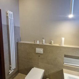 Maryan Mazan usługi budowlane - Nowoczesna łazienka z podwieszaną toaletą, beżowymi płytkami i minimalistycznym oświetleniem. Widoczny grzejnik drabinkowy i drewniany element stropu.