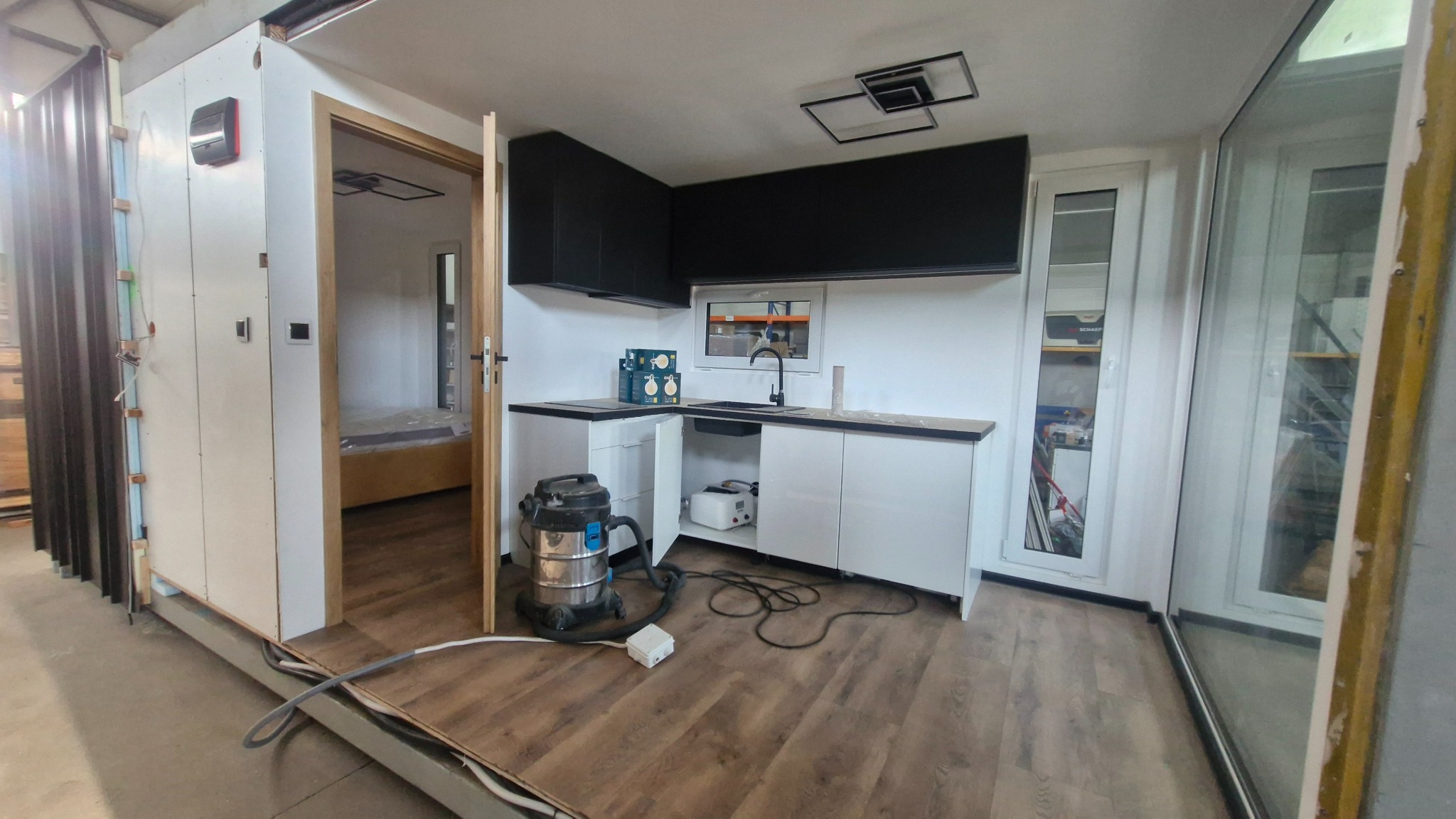 Nowoczesna kuchnia w kontenerze mieszkalnym: białe szafki, czarne górne półki, drewnopodobna podłoga, okno z widokiem na magazyn, odkurzacz na podłodze.