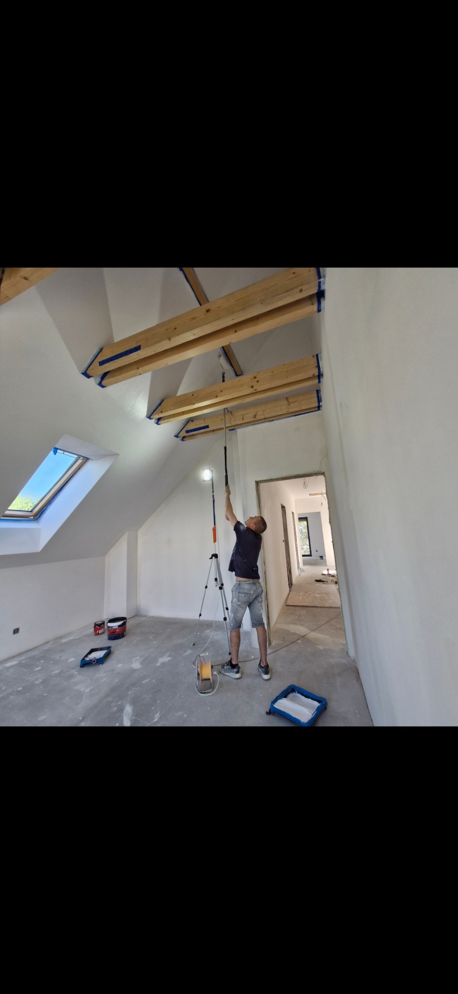 Malowanie sufitu z drewnianymi belkami w pomieszczeniu ze skosami i oknem dachowym. Widoczny malarz używający wałka na wysięgniku i lampy na statywie.