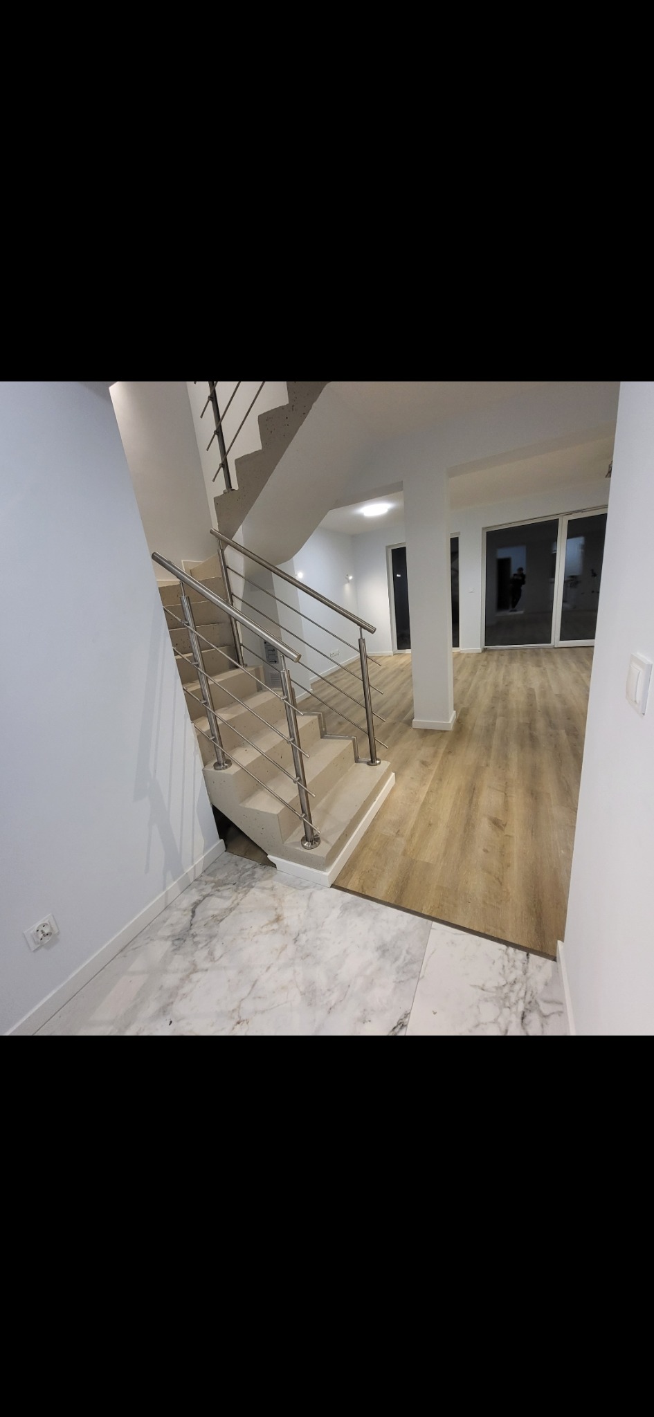 Nowoczesne schody z metalową balustradą i marmurową posadzką w przestronnym, jasnym wnętrzu. Widoczne drewniane panele podłogowe w salonie.