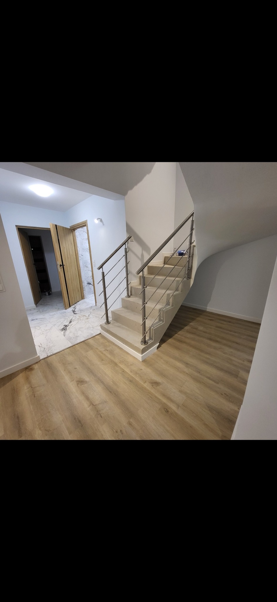 Jasne drewniane panele podłogowe i schody z metalową balustradą w nowym wnętrzu. Widoczne otwarte drzwi z ościeżnicą i marmurowa podłoga w tle. Minimalistyczny design.